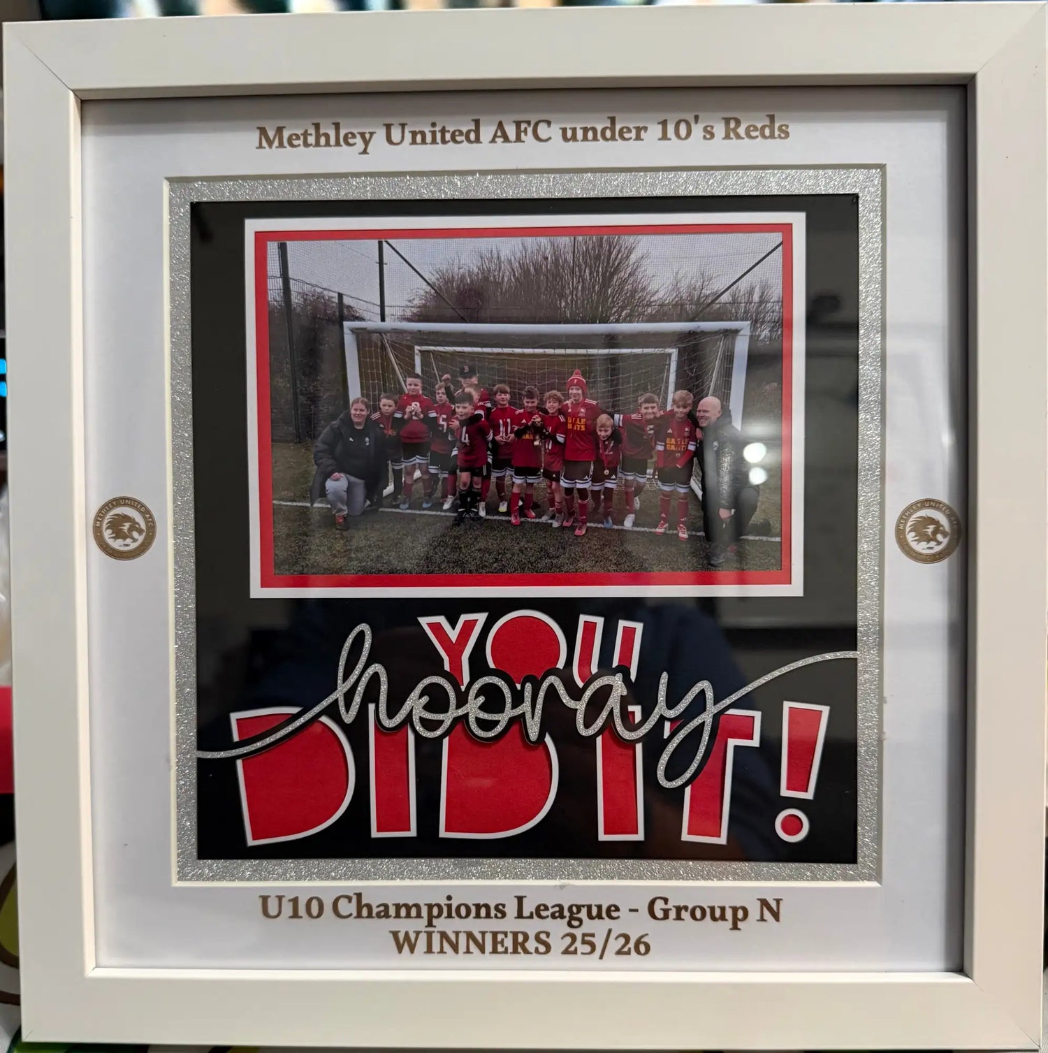 White framed sports achievement display featuring a team photo in red and black with glitter accents, celebrating the methley united afc under 10’s reds as u10 champions league group n winners.